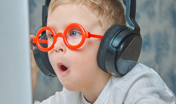 A child in red glasses and headphones has fun communicating via video link using a laptop.